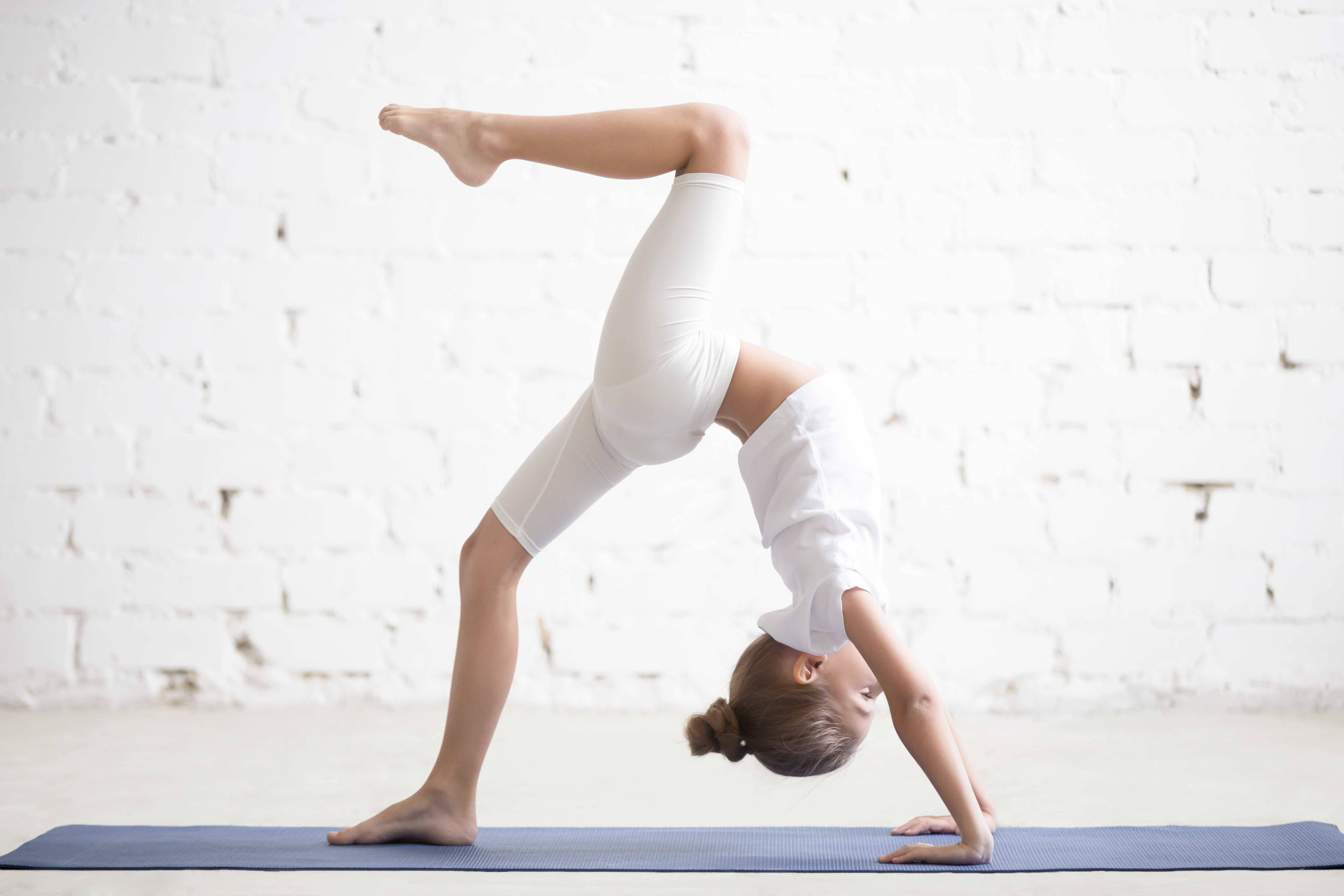 Girl child practicing yoga or fitness, standing in Bridge exercise, One legged Wheel pose, working out wearing sportswear, t-shirt, pants, indoor full length, white loft studio background